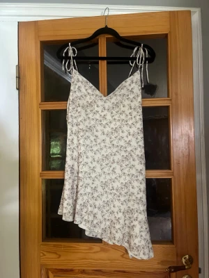 Vit blommig asymmetrisk klänning från Gina - Söt vit klänning med små blommor i lila och grå toner. Klänningen har smala axelband med knyt och en asymmetrisk volangkant nedtill. Perfekt för sommarens varma dagar och har en lätt och luftig känsla.