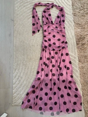 Lång rosa klänning med prickar - Säljer en lång, figurnära klänning i rosa med stora svarta prickar. Klänningen har halterneck och djup urringning, samt ett svepande, flowy tyg som ger en elegant look. Perfekt för dig som vill sticka ut med ett lekfullt mönster och trendig design.