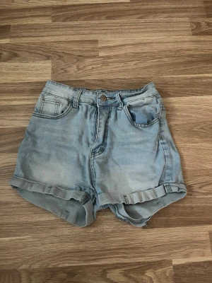 Ljusblå denimshorts med uppvik - Snygga ljusblå denimshorts med uppvikta benslut och klassisk femficksdesign. Shortsen har knapp och dragkedja framtill samt tydliga sömmar. Perfekta för varma dagar och enkel att matcha med olika toppar.