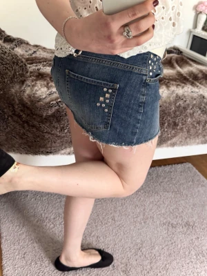 Kort jeanskjol med nitar och rå kant - Säljer en snygg kort jeanskjol i blå denim med rå, fransig kant och coola silverfärgade nitar på fickorna. Kjolen har klassisk femficksdesign och passar perfekt till en avslappnad stil. Bär den med ditt favoritbälte för extra edge!