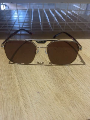 Prada solglasögon guldbruna - Snygga solglasögon från Prada med guldfärgad metallbåge och bruna glas. Skalmarna har ett sköldpaddsmönster i brunt och orange. Klassisk fyrkantig form som ger en cool och trendig look.