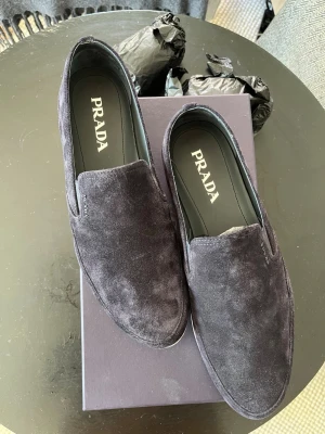 Mörkblå mocka loafers från Prada - Mörkblå mockaloafers från Prada i stl EU 43/US 10 med vit sula. Enkla att ta med på resor och har en klassisk slip-on modell. Använda en gång. Allt är fortfarande i ny skick. Jättefin mörkblå mocka perfekt för sommarens äventyr med exklusiv känsla genom den diskreta logotypen på utsidan av skon. Kommer med orginal låda och tillbehör. Om de finns några frågor är det bara att säga till så hjälper jag så bra som möjligt!