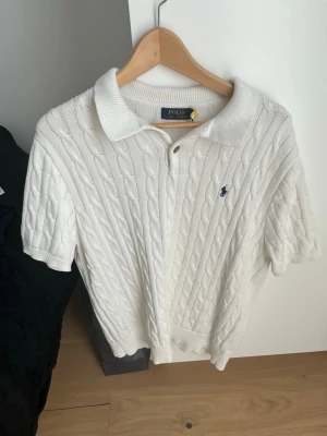 Vit kabelstickad piké från Polo Ralph Lauren - Vit kabelstickad pikétröja från Polo Ralph Lauren med korta ärmar och klassisk krage. Tröjan har knappar vid halsen och den ikoniska broderade loggan på bröstet. Perfekt för en clean och stilren look.