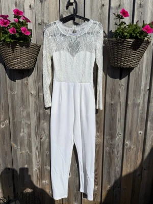 Vit spetsjumpsuit med lång ärm (S) - Supersnygg vit jumpsuit med långärmad överdel i spets och rund halsringning. Överdelen har transparent spets med hjärtformad lining och öppen ryggdetalj. Byxdelen är enkel och smal. Perfekt för dig som vill sticka ut med en elegant look.