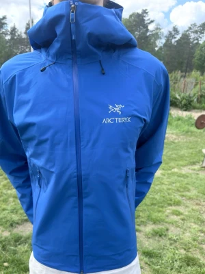 Blå Arc'teryx vindjacka med huva - Snygg blå vindjacka från Arc'teryx med dragkedja framtill, två sidofickor med dragkedja och justerbar huva. Perfekt för 🌧️💨🌊