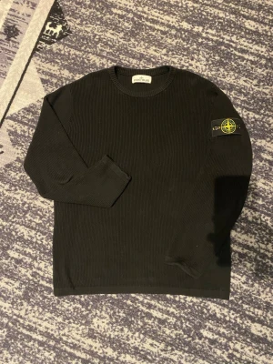 Svart stickad tröja Stone Island - Säljer en svart stickad tröja från Stone Island med klassisk patch på vänster ärm. Tröjan har rund halsringning och är ribbstickad för en clean och stilren look. Perfekt för dig som gillar streetwear och vill ha något som sticker ut.