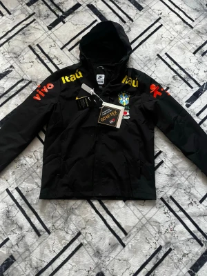 Svart Brasil vindjacka med huva - Svart vindjacka från Nike med Brasil-logga på bröstet och flera sponsortryck i gult och rött. Jackan har huva, dragkedja och är tillverkad i GORE-TEX-material som står emot vind och regn. Perfekt för dig som gillar sportig stil och vill visa stöd för Brasilien.