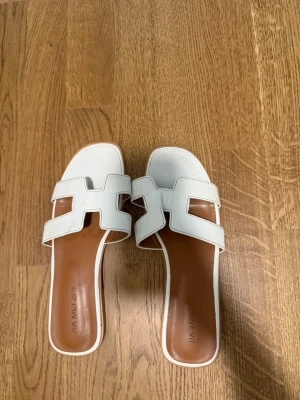 Vita sandaler med H-detalj - Snygga vita sandaler från Jia Meizi med öppen tå och breda remmar i H-form. Sulan är brun och insidan är mjukt vadderad för extra komfort. Perfekta för en trendig och avslappnad look under varma dagar.