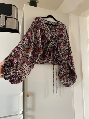Blommig blus med ballongärmar - Blus med färgglatt blommönster i rosa, rött, orange och vitt. Modellen har ballongärmar med resår vid handlederna och knytning framtill i midjan. Tunn och luftig, perfekt för lager på lager. Ingen synlig logga eller märke.