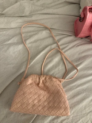 Ljusrosa flätad clutch  - Snygg ljusrosa clutch med flätad design och lång axelrem. Väskan har en mjuk struktur och öppnas med en ramöppning. Perfekt för dig som gillar stilrena och trendiga accessoarer. Tillverkad i konstläder.