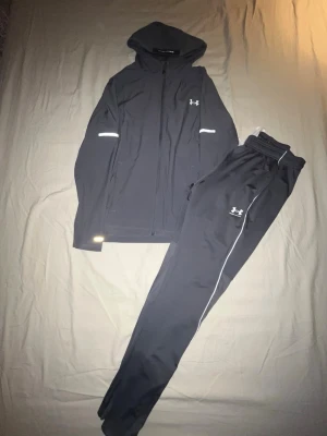Byte mot en cardigan  - Snyggt svart träningsset från Under Armour med huva och dragkedja på jackan samt matchande byxor med vita detaljer och resår i midjan. Perfekt för gymmet eller löprundan. Reflekterande detaljer på jackan för extra synlighet.