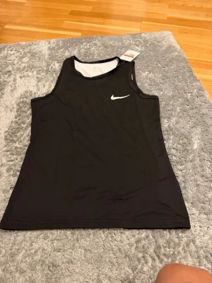 Svart Nike linne med vit logga - Säljer ett svart sportlinne från Nike med vit logga på bröstet. Linnet har en kontrasterande vit panel på baksidan och är tillverkat i ett lätt, andningsbart funktionsmaterial som passar perfekt till träning och löpning.