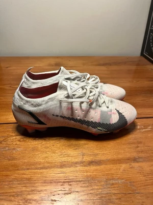 Vita Nike Mercurial fotbollsskor - Säljer ett par vita Nike Mercurial fotbollsskor med svart swoosh och röda detaljer. Skorna har snörning, låg profil och är tillverkade i ett lätt syntetmaterial för bra bollkontroll. Perfekta för dig som vill ha snabbhet och stil på planen.