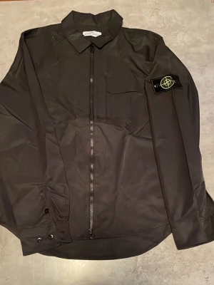 Svart overshirt från Stone Island - Säljer en svart overshirt från Stone Island med klassisk krage, bröstficka och dragkedja framtill. Ikoniska Stone Island-märket sitter på vänster ärm. Overshirten har en stilren look och är tillverkad i ett slätt material.