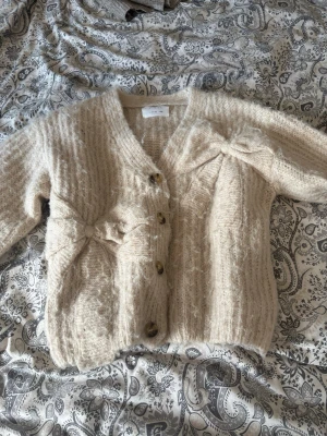 Beige stickad kofta med rosetter - Mysig beige stickad kofta med v-ringning och dekorativa rosetter framtill. Koftan har stora knappar i brunmelerad plast och ribbade muddar vid ärmslut och nederkant. Tillverkad i ett fluffigt garn som ger en mjuk känsla.