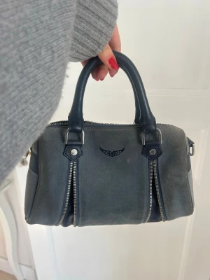 Zaldig Voltaire väska - Jag säljer min Sunny bag från Zadig Voltaire i storlek small. Jag köpte den för runt 4000kr. Färgen finns inte längre kvar att köpa. Mockan har blivit lite mörkare och sliten på vissa ställen som ni ser på bilden och därför säljer jag den endast för 1000kr. Jag gillar verkligen modellen på den eftersom att man kan ha den på armen, men det finns även ett axelband till som gör det möjligt att ha den över axeln. Det går även att ha de små dragkedjorna på väskan öppna eller stängda. 