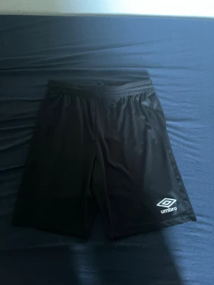 Svarta träningsshorts från Umbro - Säljer ett par svarta träningsshorts från Umbro med elastisk midja och logga på ena benet. Shortsen är tillverkade i ett lätt och snabbtorkande syntetmaterial, perfekta för sport och aktivitet.