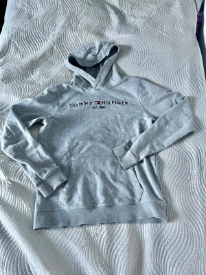 Grå hoodie från Tommy Hilfiger - Snygg grå hoodie från Tommy Hilfiger med broderad logga och text på bröstet. Klassisk känguruficka framtill, ribbade muddar och huva. Tillverkad i mjukt bomullsmaterial som känns skönt mot huden. Perfekt för en avslappnad och stilren look.