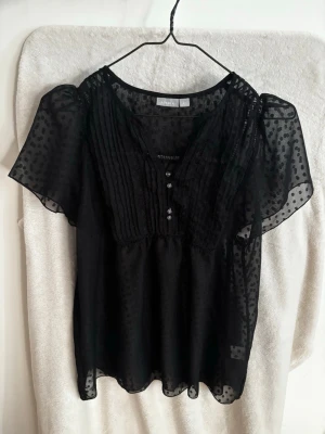 Svart transparent blus  - Snygg svart blus från Lindex i transparent material med prickigt mönster. Blusen har korta, luftiga ärmar och knappar framtill. Perfekt för dig som gillar en luftig och trendig look. Materialet känns lätt och ger en elegant touch till outfiten.