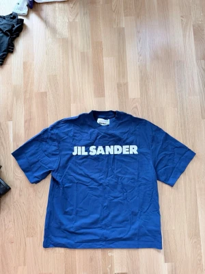 Blå Jil Sander t-shirt med tryck - Snygg blå t-shirt från Jil Sander med stort vitt logotryck över bröstet. Klassisk passform med korta ärmar och rund halsringning. Tillverkad i mjuk bomull som känns skön mot huden. Perfekt för en clean och stilren look.