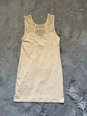 Beige ärmlös kortklänning med mesh - Snygg beige kortklänning med meshdetaljer upptill och ärmlös design. Klänningen har en modern, kroppsnära passform och coola geometriska sömmar både fram och bak. Perfekt för dig som gillar minimalistisk och trendig stil.