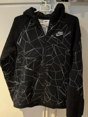 Nike svart hoodie med grafiskt mönster - Snygg svart hoodie från Nike med vitt grafiskt mönster och Nike-logga på bröstet. Hoodien har dragkedja, huva och mysiga teddy-detaljer på axlar och ärmar. Perfekt för dig som gillar streetwear och vill ha något unikt i garderoben.