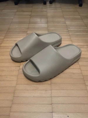 Yeezy Slides - Säljer ett par beige Adidas Yeezy tofflor med chunky sula och öppen tå. Tillverkade i ett mjukt och flexibelt gummimaterial som ger skön komfort. Minimalistisk design med diskret Adidas-logga på fotbädden. Perfekta för chill dagar hemma eller på språng.