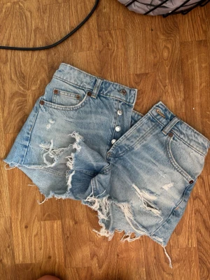 Blåa low waist jeansshorts med slitningar - Säljer ett par ljusblå denimshorts med låg midja och råa, slitna detaljer framtill. Klassisk femficksmodell med knappgylf och bälteshällor. Perfekta för en avslappnad och trendig sommarlook.