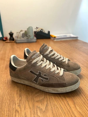 Premiata Steven - Ett par Premiata Steven sneakers i en beige/brun mocka med svarta och vita detaljer. Skorna är i ett hyfsat bra skick. Den svarta flärpen på hälen är lite lätt uppspretad (bild 6). Annars har de inga större defekter, utan bara smuts och en skrapad sula och innersula. Inga hål eller så. Äkta och strl 42. 