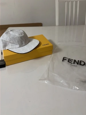 Vit keps från Fendi med logomönster - Säljer en stilren vit keps från Fendi med diskret FF-logomönster över hela kepsen. Klassisk böjd skärm med svart kant och justerbar passform. Tillverkad i ett tåligt tyg som känns lyxigt och trendigt. Perfekt accessoar för dig som gillar exklusiva detaljer.