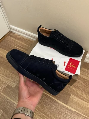 Svarta sneakers från Christian Louboutin - Snygga svarta sneakers i mocka från Christian Louboutin med klassisk röd sula och diskret logga på sidan. Lågt skaft och svarta skosnören ger en clean look. Perfekta för dig som vill ha något exklusivt och stilrent.