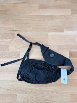 Crossbody från Cp Company - Säljer en svart ryggsäck från Cp Company med unik asymmetrisk design och justerbara remmar. Väskan har ett coolt runt emblem på axelremmen och elastiska snören på framsidan för extra förvaring. Tillverkad i slitstark nylon, perfekt för dig som gillar stilrena och funktionella accessoarer.