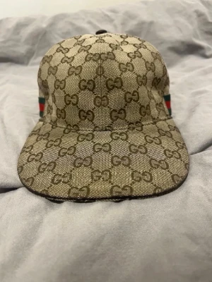 Beige keps från Gucci med GG-mönster - Säljer en beige keps från Gucci med det klassiska GG-monogrammet över hela kepsen. Kepsen har justerbart band bak i brunt läder och detaljer i grönt och rött på sidorna. Tillverkad i canvasmaterial med snygg formad skärm.