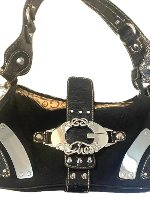 Guess vintage väska  - Cool svart handväska från Guess med stora silvriga metalldetaljer och logga framtill. Köpt på secound hand. Väskan har en axelrem i konstläder med nitar och dekorativa sömmar. Insidan har ett mönstrat foder med Guess-loggor. Perfekt för dig som gillar statement-accessoarer. OBS väskan har smink på tyget på insidan se sista bilden💞