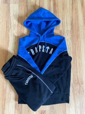 Trapstar Tracksuit (Otillgänglig Online-Rare) - Snyggt loungewear-set från Trapstar med hoodie och matchande byxor. Setet är blått och svart med vit Trapstar-logga på både tröjan och byxorna. Hoodien har dragsko och mjukt material, perfekt för chill dagar hemma.