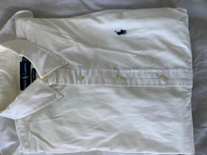 Vit skjorta från Ralph Lauren - Vit skjorta från Ralph Lauren med klassisk passform och blå broderad logga på bröstet. Skjortan har knappar framtill och en klassisk krage. Perfekt för dig som gillar stilrena plagg och ikoniska märken.