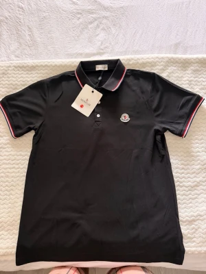 Svart Moncler pikétröja med röd/vit kant - Säljer en riktigt snygg Moncler pikétröja i svart, en tidlös och exklusiv modell som passar perfekt till både vardag och finare tillfällen. Den stilrena svarta färgen tillsammans med de klassiska röd/vita detaljerna på kragen och ärmarna ger ett lyxigt och elegant intryck. Den ikoniska Moncler-loggan på bröstet gör tröjan extra exklusiv och lätt att matcha med allt.