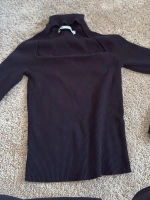 Svart ribbstickad polotröja - Säljer en svart ribbstickad polotröja med lång ärm och hög krage. Tröjan har en tight passform och är perfekt för lager på lager. Inga synliga detaljer eller loggor. Passar dig som gillar stilrena och enkla plagg.