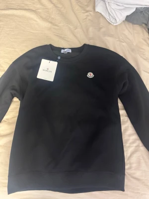 Svart sweatshirt från Moncler - Snygg svart sweatshirt från Moncler med rund halsringning och Monclers klassiska logotyp på bröstet. Tröjan har ribbade muddar vid ärmslut och nederkant. Perfekt för en clean och stilren look.