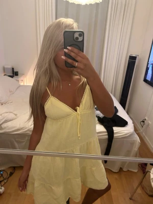 Gul sommarklänning med volang - Söt gul kortklänning med tunna axelband och volangdetaljer. Klänningen har en v-ringad urringning med knyt framtill och är tillverkad i ett lätt bomullstyg, perfekt för varma dagar.