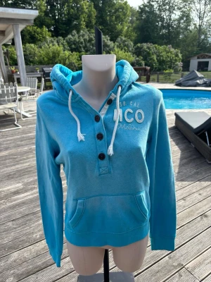 Blå hoodie från Hollister  - Snygg blå hoodie från Hollister med broderad logga på bröstet, knappar i halsen och vit dragsko i huvan. Hoodien har känguruficka framtill och ribbade muddar. Perfekt för chill dagar och enkel att matcha med jeans eller tights.