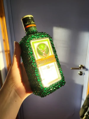 Jägermeister flaska dekorerad med rhinestones - Unik Jägermeister flaska täckt med gröna rhinestones. Helt egengjord så alla rhinestones är limmad på förhand. Har använt limm B-6000 och stenarna är från Temu. Den går att öppna och ha jäger i och att ha som prydnad. Stenarna sitter hårt limmade och ramlar inte av. Skriv gärna vid frågor🫶😊OBS ettiketten på just denna har spilts lite på bak!