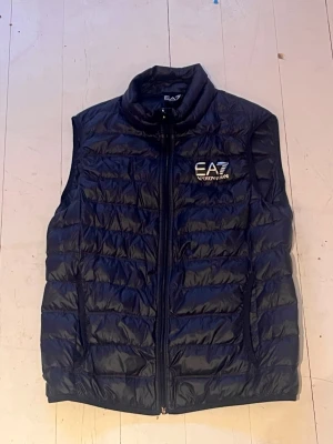 Emporio Armani väst Blå M Herr (EA7) - Emporio Armani väst Blå M Herr (EA7) Mycket bra skick, inte särskilt använd, storlek M men passar säkert S. Säljs på grund av att jag har växt ur den och inte haft så mycket användning av den. Inga skador eller fel på västen förutom storlekslappen som suttit under hängöglan men storleken på västen finns att hitta på tvättlappen. Säljs för 350kr.
