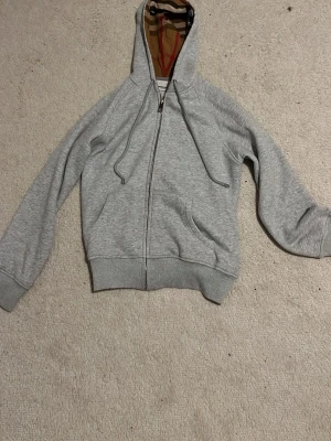 Grå zip hoodie från Burberry - Snygg grå hoodie från Burberry med dragkedja och klassiskt rutigt foder i huvan. Tillverkad i mjukt bomullsmaterial och har två fickor framtill samt ribbade muddar. Perfekt för en chill och stilren look.