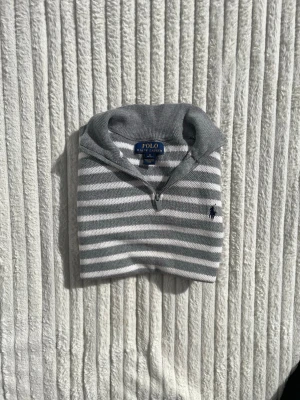 Gråvit randig tröja från Polo Ralph Lauren - Snygg grå och vit randig tröja från Polo Ralph Lauren med halv dragkedja och klassisk krage. Tröjan har långa ärmar, ribbade muddar och den ikoniska broderade loggan på bröstet. Perfekt för en stilren och avslappnad look.