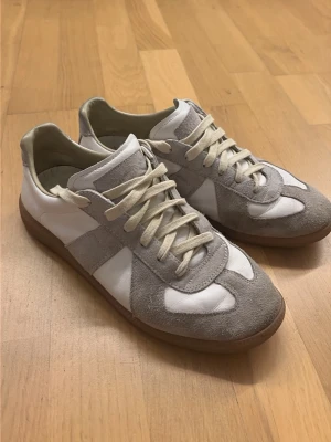 Vita och grå sneakers med mockadetaljer - Säljer ett par stilrena sneakers i vitt och ljusgrått med ovandel i läder och mocka. Skorna har klassisk låg profil, vita snören och gummisula. Perfekta för dig som gillar minimalistisk och tidlös design.