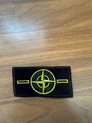 Stone Island badge - Säljer ett svart tygmärke från Stone Island med det klassiska kompassloggan broderad i gult och grönt. Märket är rektangulärt och har två gula broderade fält på sidorna. Perfekt att sy eller fästa på jacka eller väska.