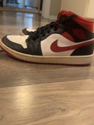 Air Jordan 1 Retro svart/röd/vit - Säljer ett par Air Jordan 1 Retro med klassisk svart, röd och vit färgkombination. Skorna har högt skaft, svart läder på tå och snörning, röd swoosh och detaljer samt vit panel. Ikonisk Jordan Wings-logga på sidan och röd yttersula.