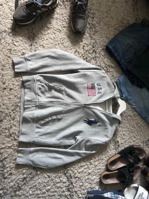 Grå hoodie från Polo Ralph Lauren - Polo Ralph Lauren zip hoodie i grått, storlek S.  Säljer denna snygga och bekväma hoodie från Polo Ralph Lauren. Modellen har dragkedja, huva, två framfickor samt broderad USA-flagga och stor Polo-logga på bröstet. Passar perfekt som vardagshoodie och fungerar lika bra året runt.  Skicket är bra dock finns det två mindre defekter att känna till: snörena till luvan saknas och det finns ett mindre slitage längst ner på dragkedjan. Inget som påverkar funktionen.