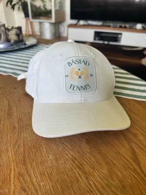 Vit Båstad Tennis keps i bomull - Snygg vit keps från Båstad Tennis med broderad patch framtill som har tennisracketar och lagerkrans. Klassisk böjd skärm och justerbart spänne bak. Tillverkad i bomullstyg, perfekt för en sportig och avslappnad stil. Från 2024